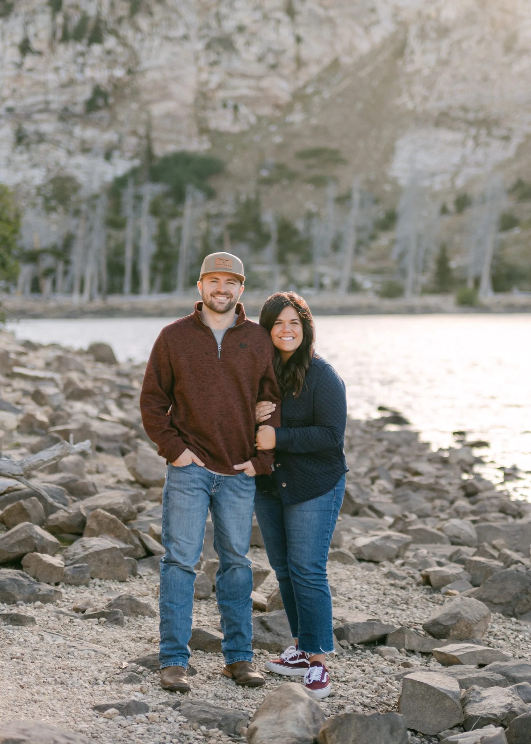 Craig and Nicole smiling together on a rocky lakeshore with mountains behind them